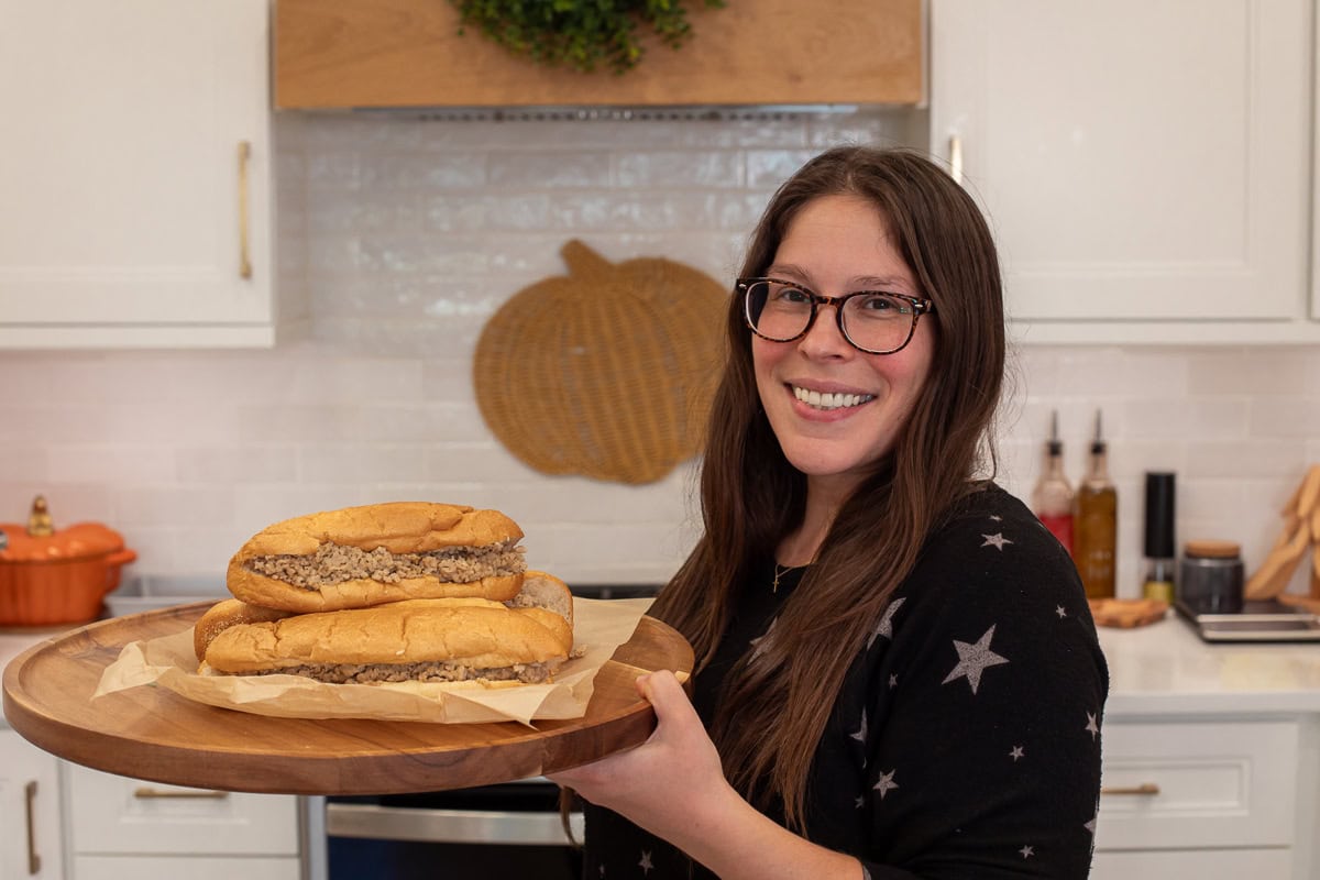 Smiling woman in a kitchen holding a wooden platter with chicken cheesesteaks on Amoroso rolls filled with ground chicken and Cooper Sharp cheese.