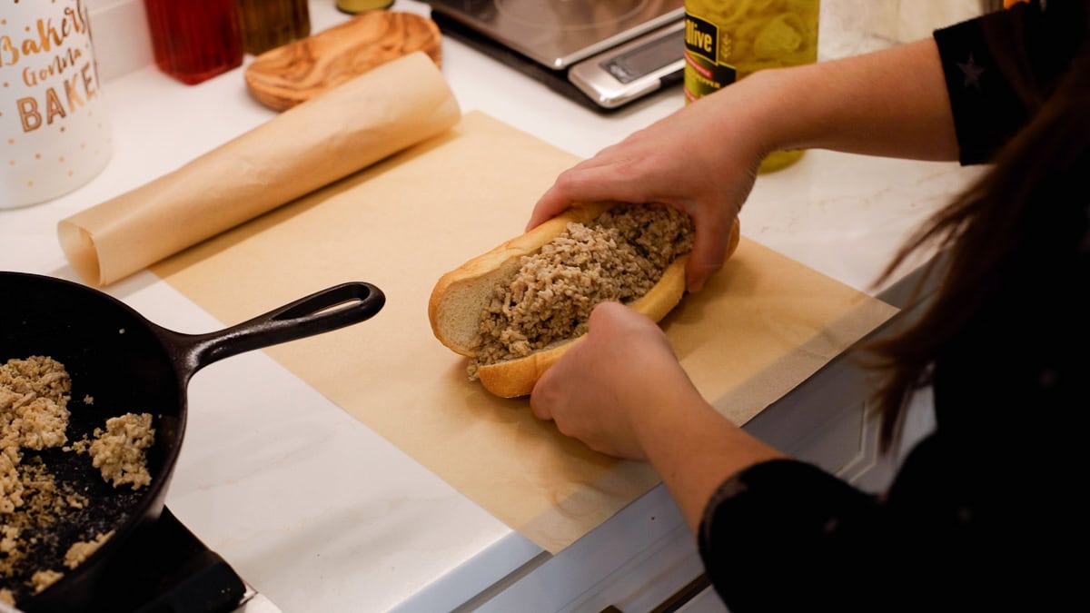 Hands holding a chicken cheesesteak placing it on parchment paper to tightly wrap it with a warm skillet to the side.