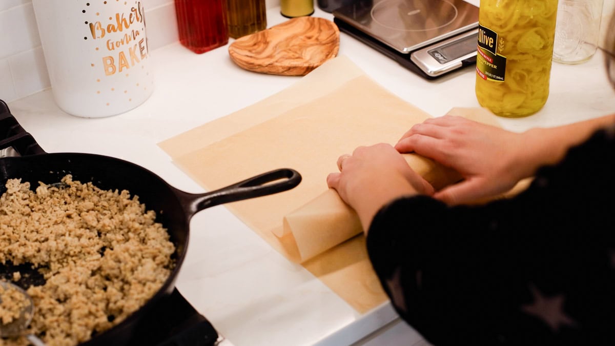 Hands rolling parchment paper to tightly wrap a chicken cheesesteak sandwich, with cooked ground chicken in a cast iron skillet nearby