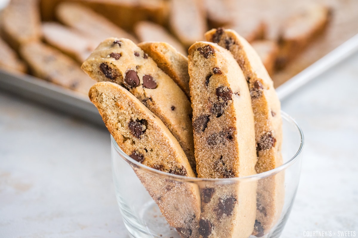 close up of sliced up mandel bread pieces in a glass bowl with mandel bread sliced on a sheet pan in the background