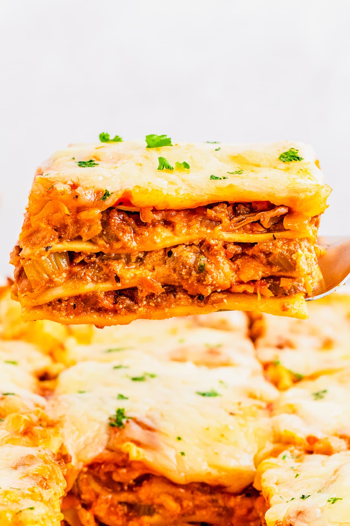 lasagna bolognese on a spatula being held up in the air above baking dish.
