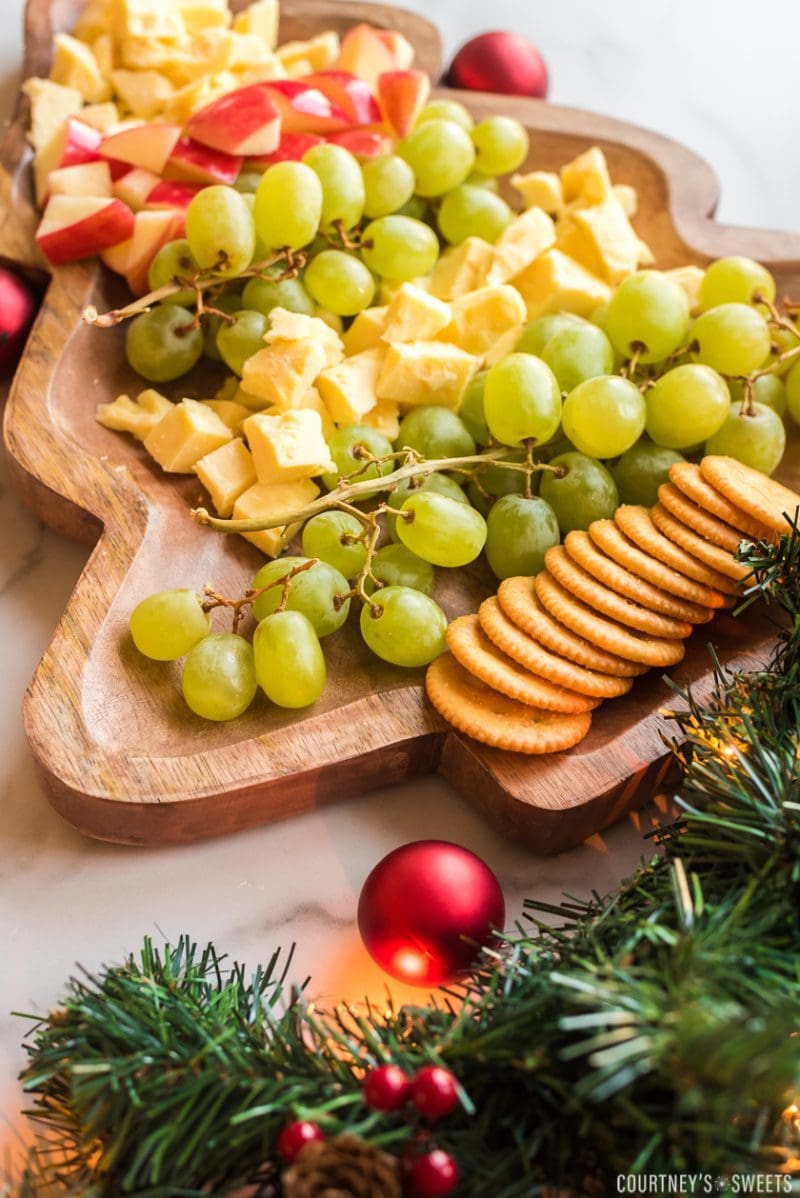 christmas tree shaped board with fruit and cheese on it with crackers and garland decor on the bottom