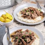 mississippi pot roast on two gray plates over mashed potatoes with peppers in a white bowl and slow cooker to the top left