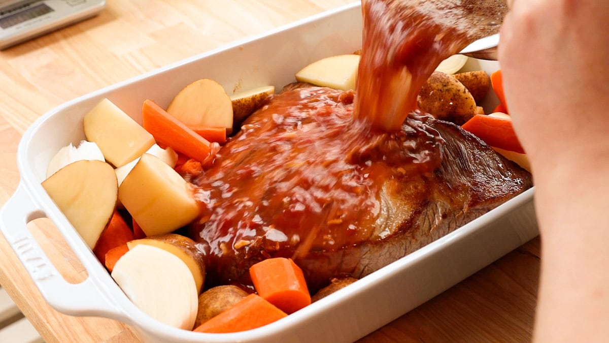 pouring sauce over brisket in a casserole dish.