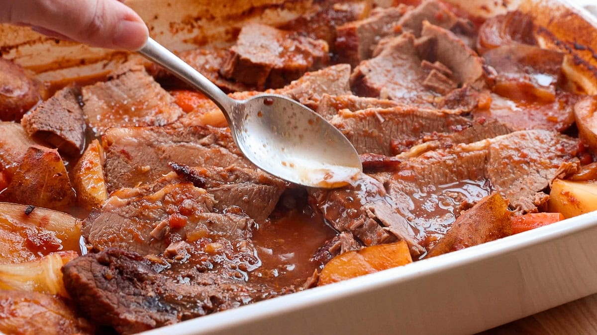spooning sauce over brisket in a casserole dish.