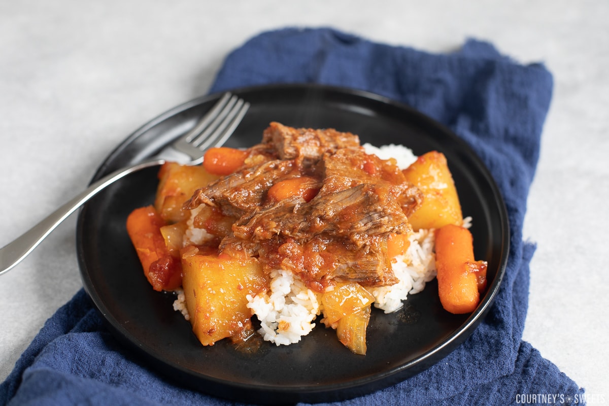 sweet brisket with carrots and potatoes over white rice on black plate with navy blue napkin underneath