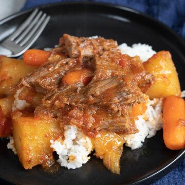 passover brisket with carrots and potatoes over white rice on black plate with navy blue napkin underneath