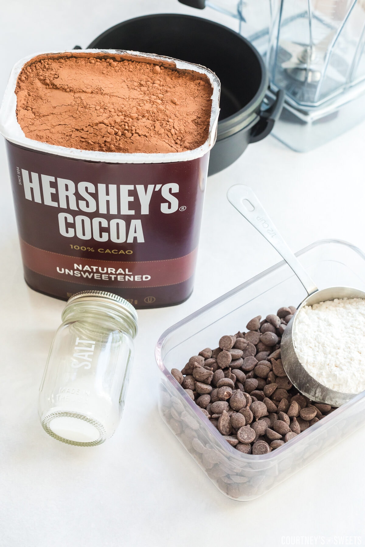 hershey cocoa powder, salt container, chocolate chips in a storage container and a measuring cup filled with powdered sugar on a table