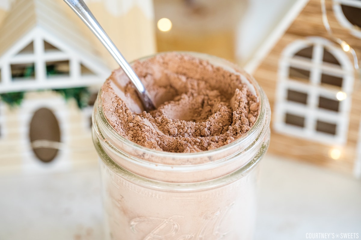 dairy free hot chocolate mix in a mason jar with spoon inside of it and little christmas houses behind it