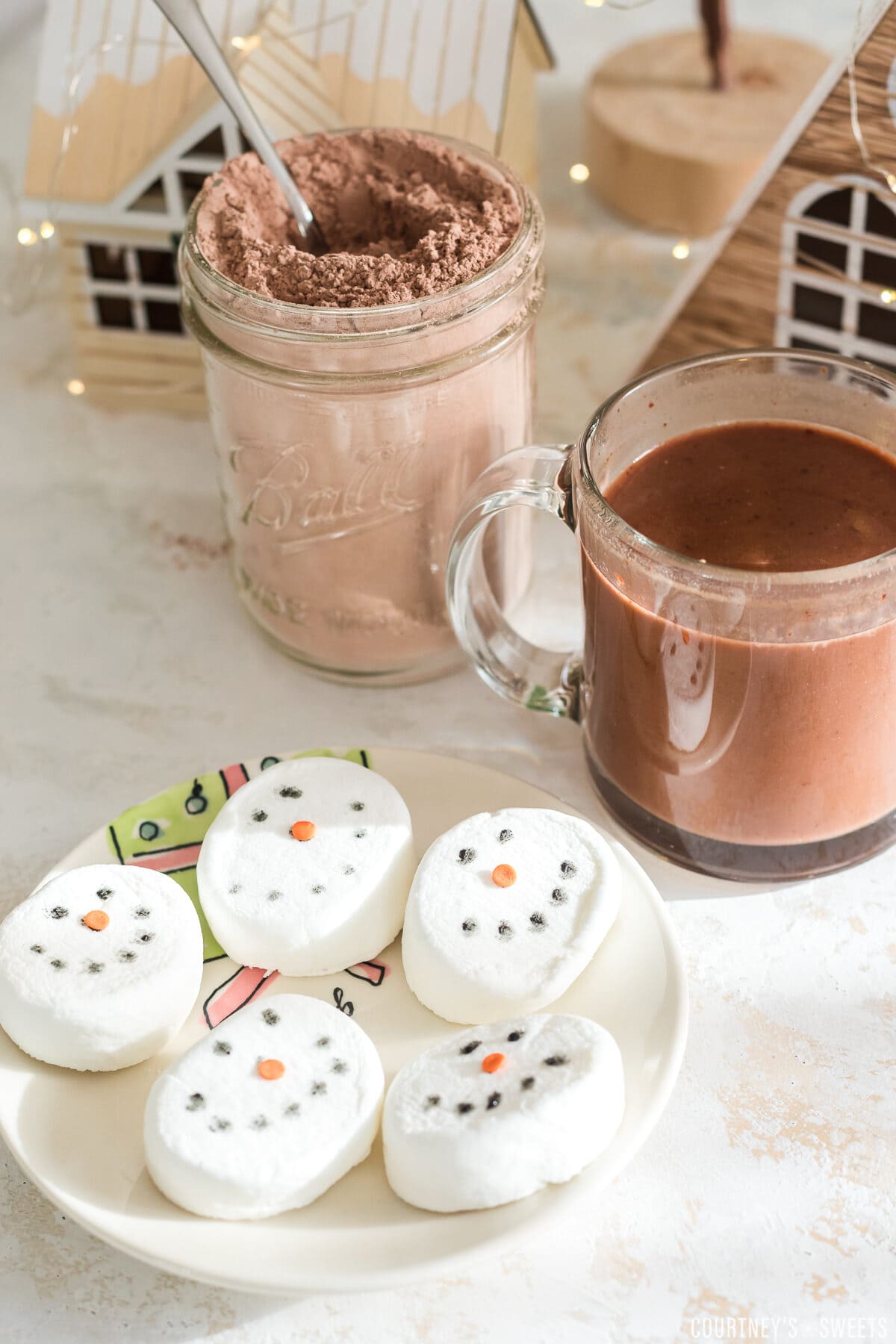 snowman marshmallow toppers on a plate with hot chocolate in a glass mug behind it and a mason jar with hot chocolate mix