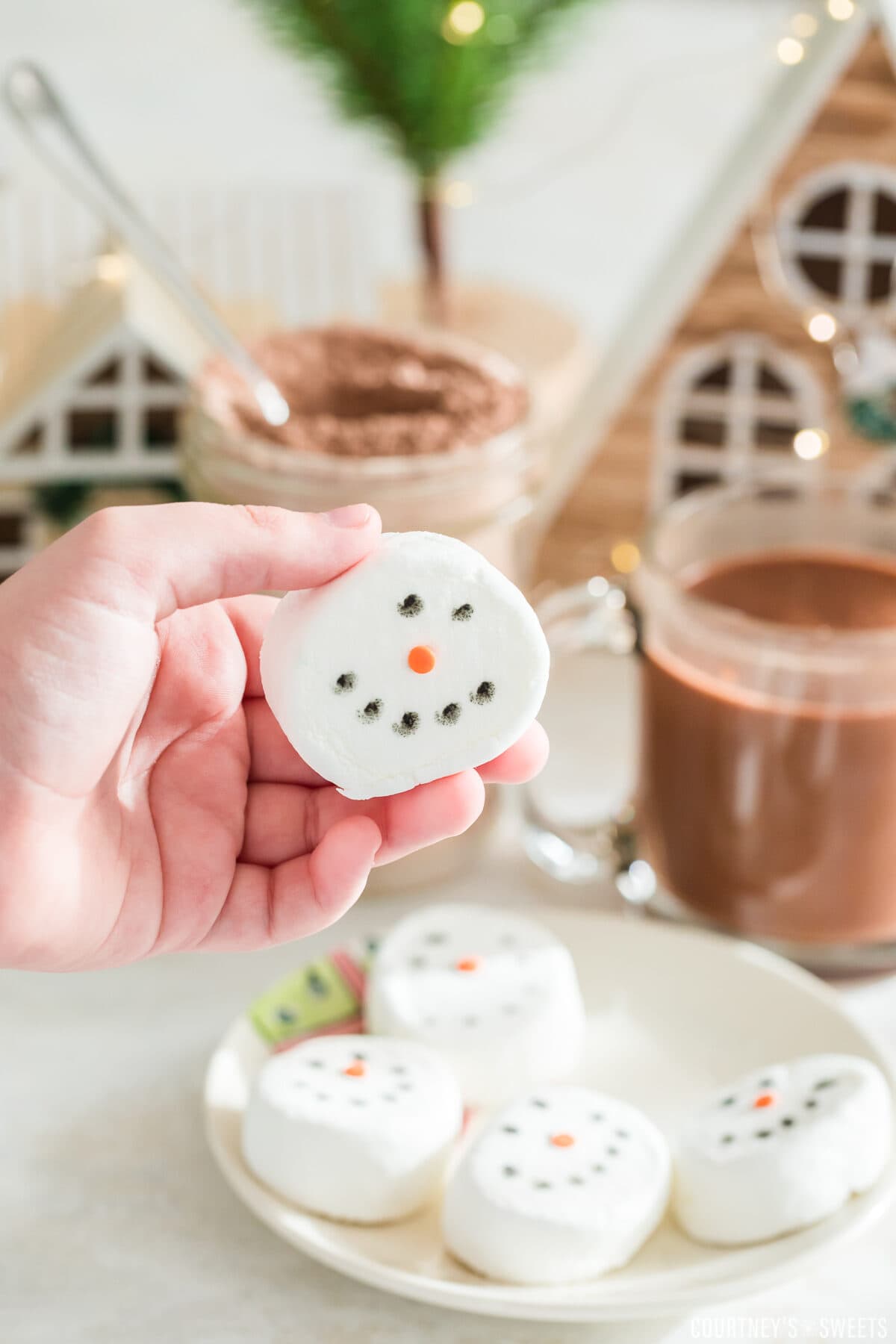 child hand holding up marshmallow snowman with plate of them under the hand and hot chocolate in the background with little christmas houses