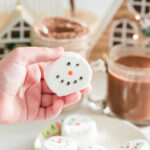 child hand holding up marshmallow snowman with plate of them under the hand and hot chocolate in the background with little christmas houses