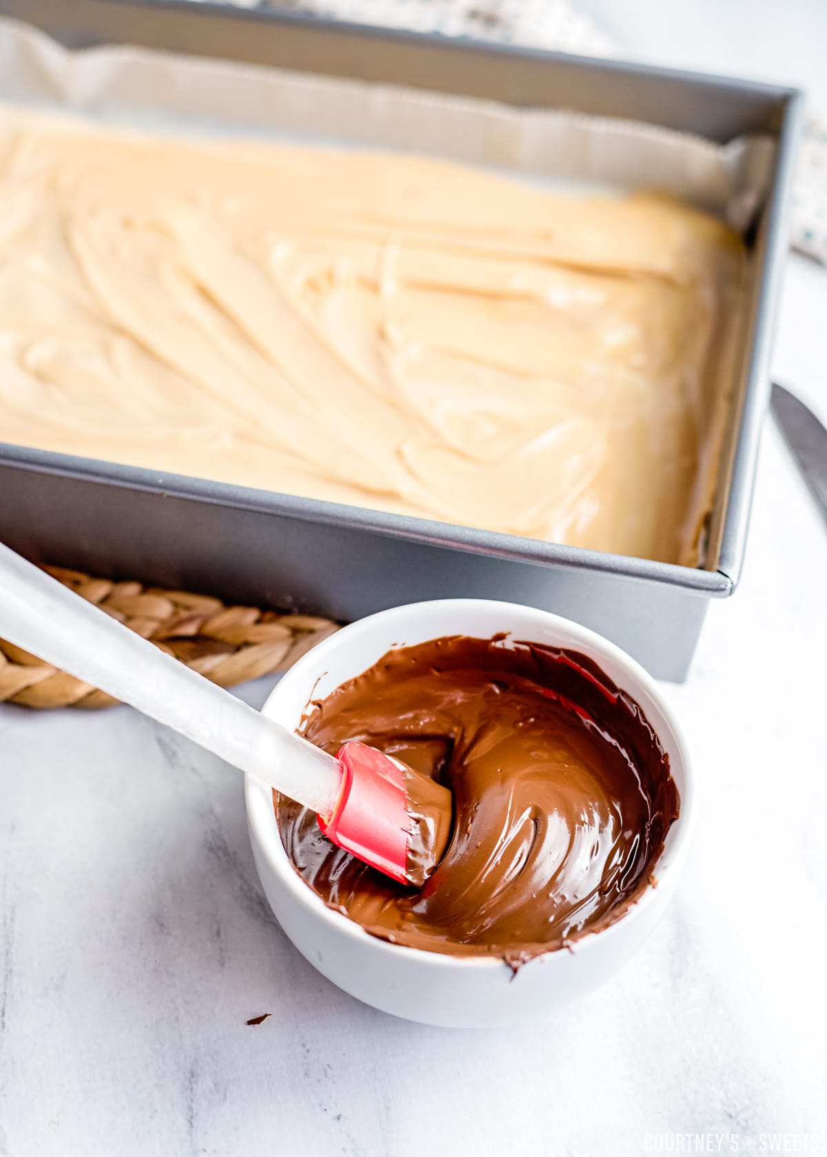 melted chocolate in a bowl with spatula and melted white chocolate and peanut butter mixed in baking dish behind it