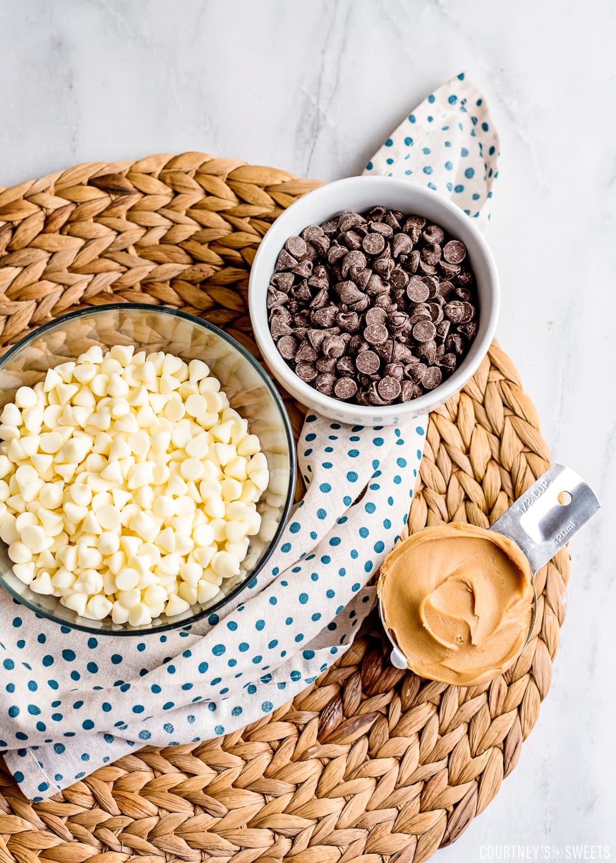 white chocolate in a glass bowl, chocolate chips in a white bowl, and peanut butter in a measuring cup on a napkin and wicker placemat