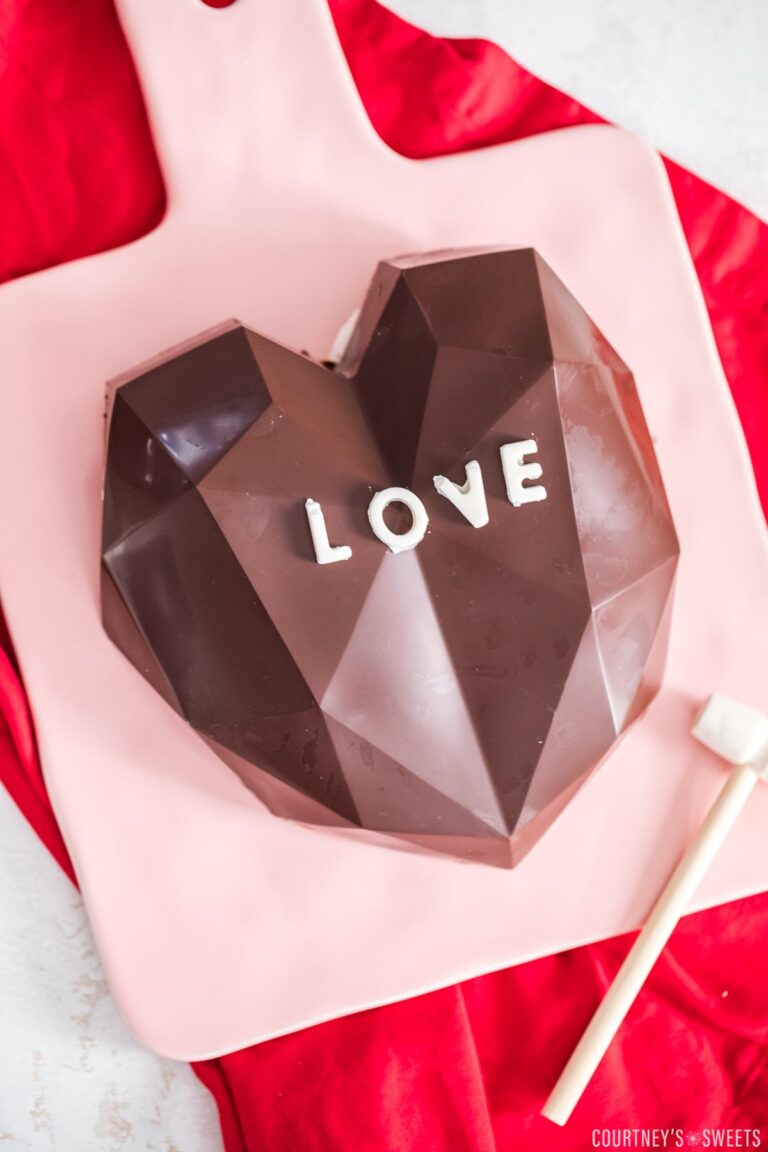 breakable chocolate heart on a pink cutting board with a wooden hammer next to it.
