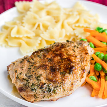 pan fried pork chops with carrots peas and buttered bowties on a white plate.