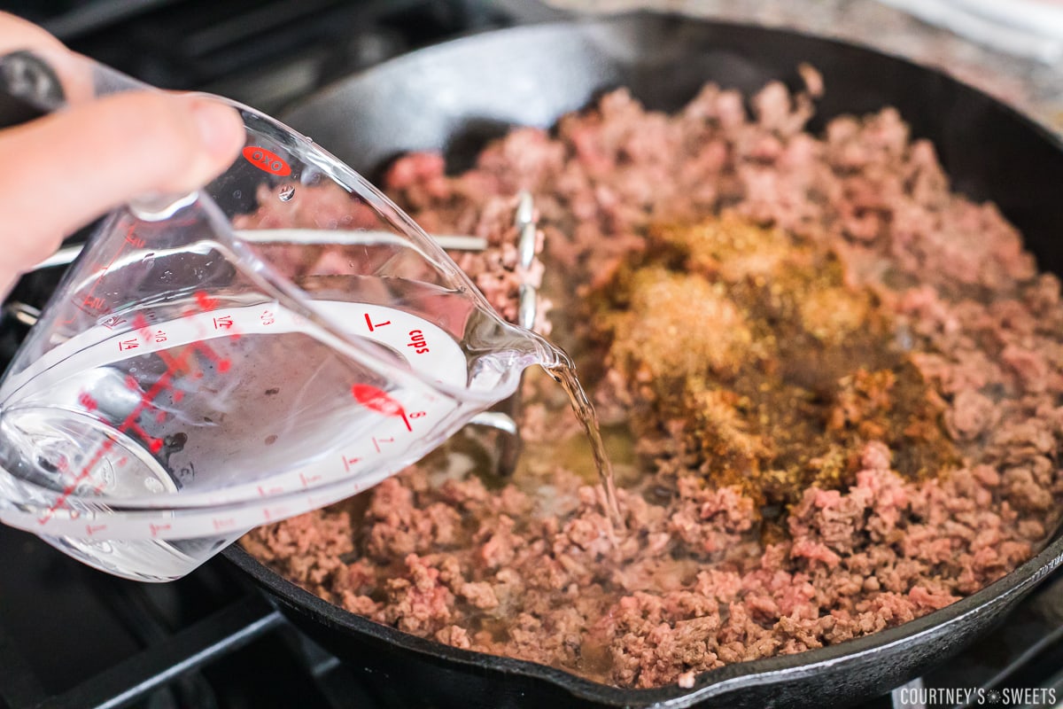 hand pouring water with measuring cup into ground beef in cast iron skillet.