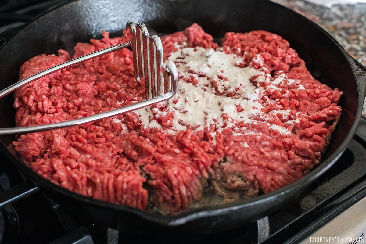 ground beef in a cast iron skillet with potato masher and flour on meat.