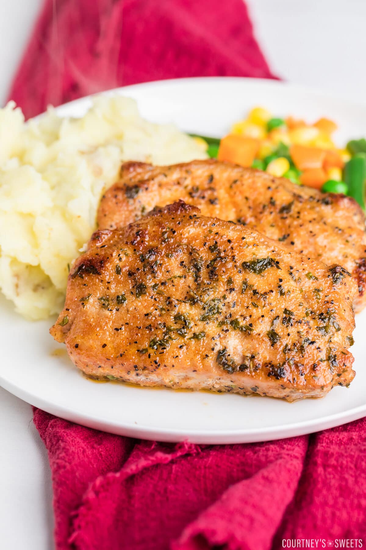 2 baked boneless pork chops on a white plate with mixed vegetables and mashed potatoes on a maroon napkin.