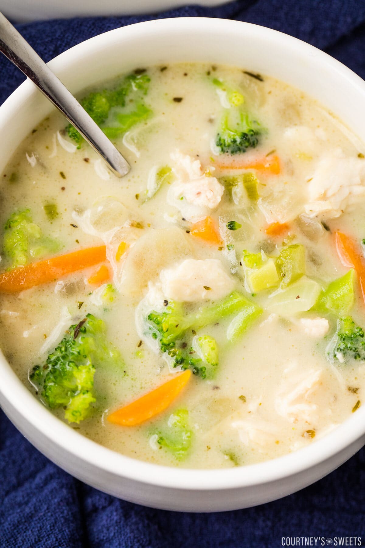 close up photo of chicken broccoli soup in a white bowl with a spoon in it on a navy blue napkin.