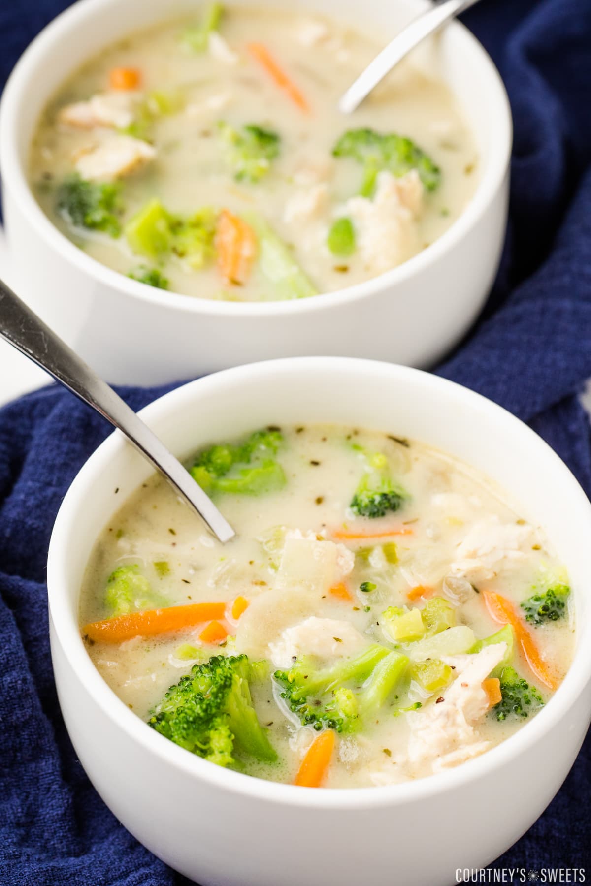 two small bowls of chicken broccoli soup with a spoon on a navy blue cloth napkin.