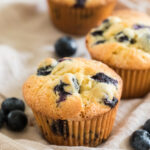 3 blueberry muffins on a beige napkin with blueberries scattered.