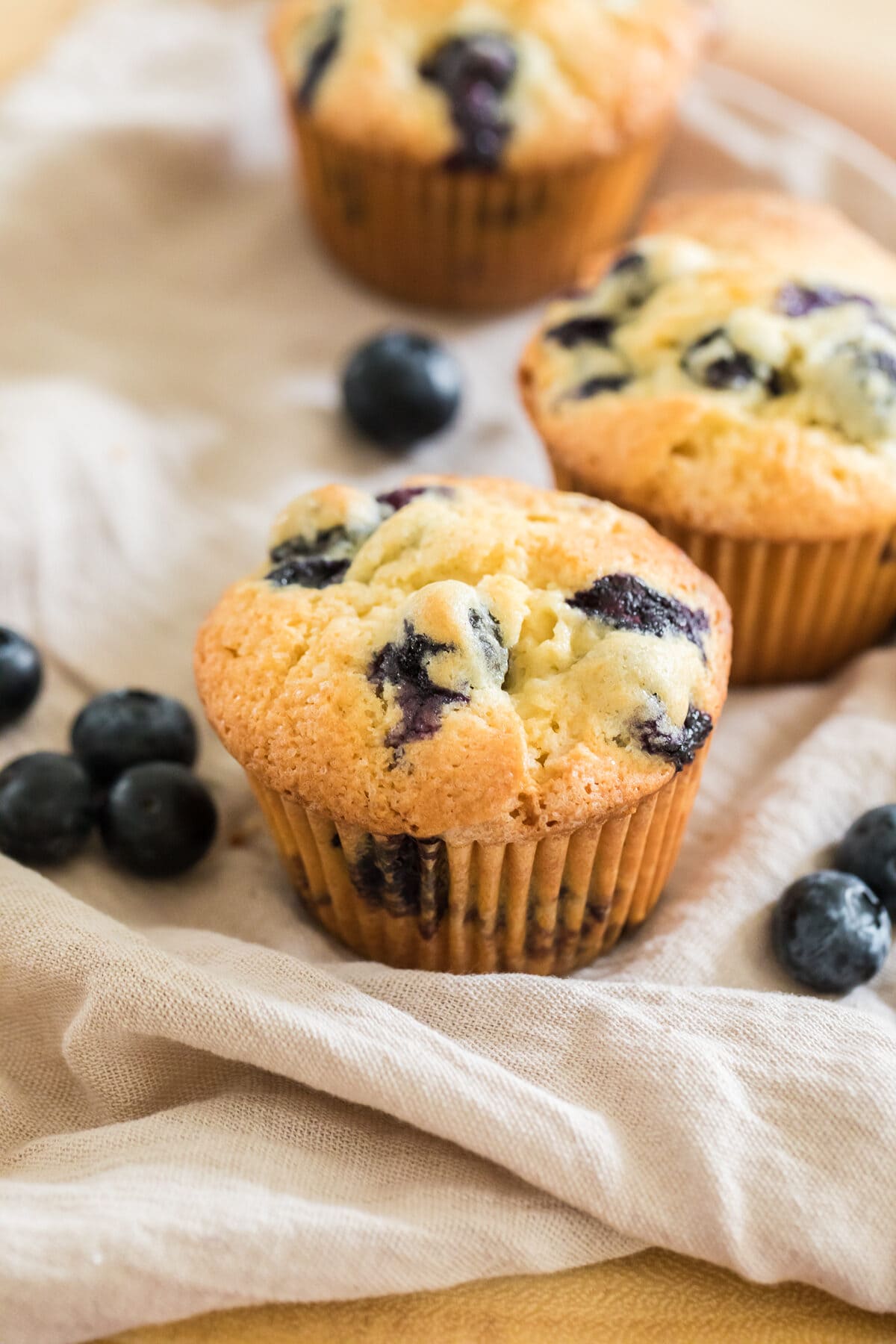 homemade blueberry muffins on a beige napkin with blueberries scattered.