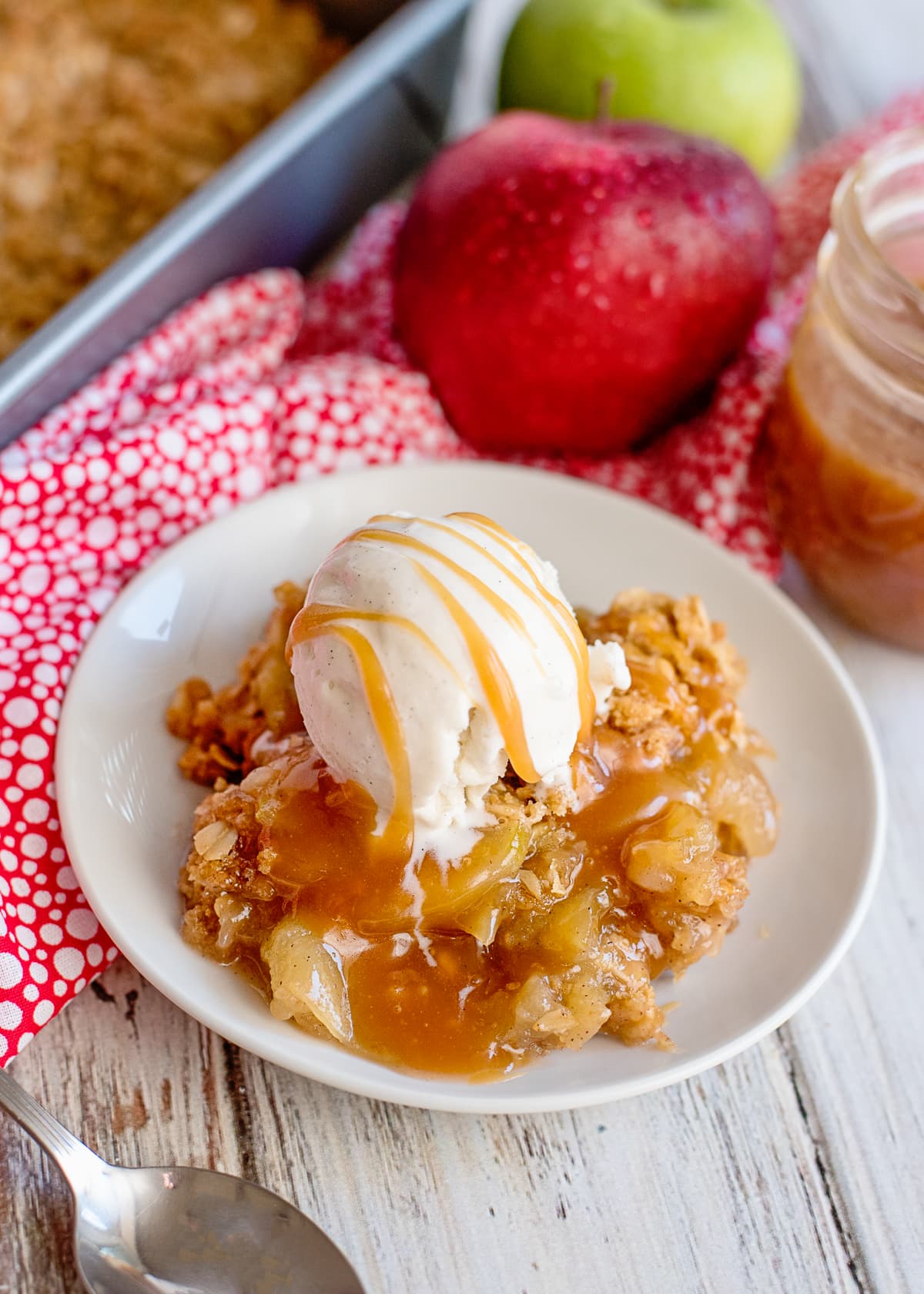 apple crisp with a scoop of vanilla ice cream and a drizzle of caramel sauce on a white plate with apple in the background.