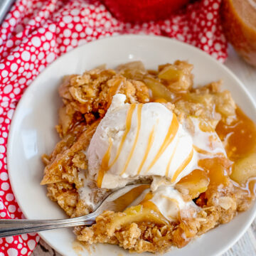 apple crisp with a scoop of vanilla ice cream and a drizzle of caramel sauce on a white plate with a spoon digging in.