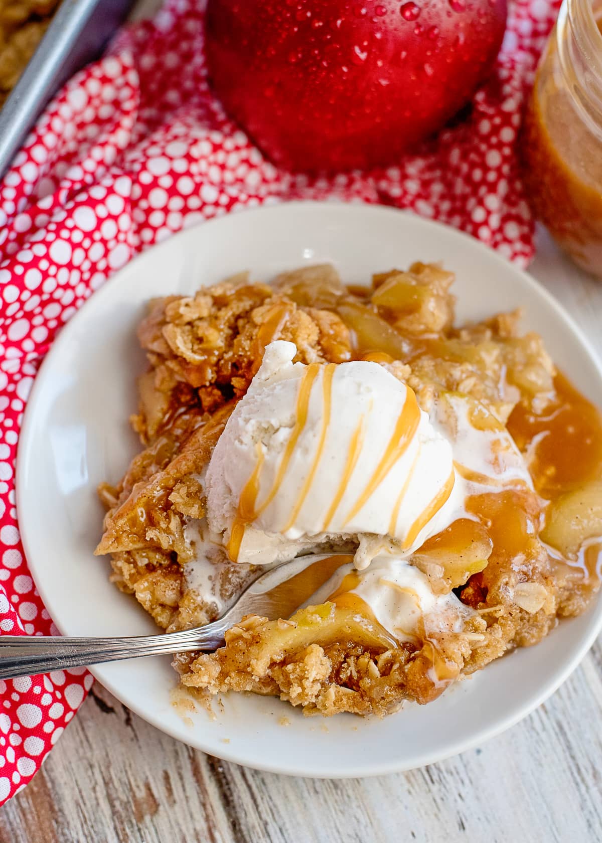 apple crisp with a scoop of vanilla ice cream and a drizzle of caramel sauce on a white plate with a spoon digging in.