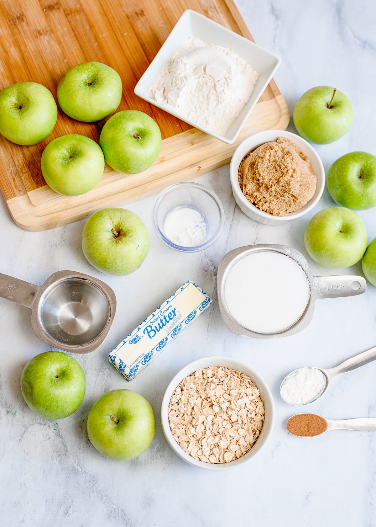 apples, stick of butter, and other baking ingredients in measuring cups for a recipe on a marble slab.
