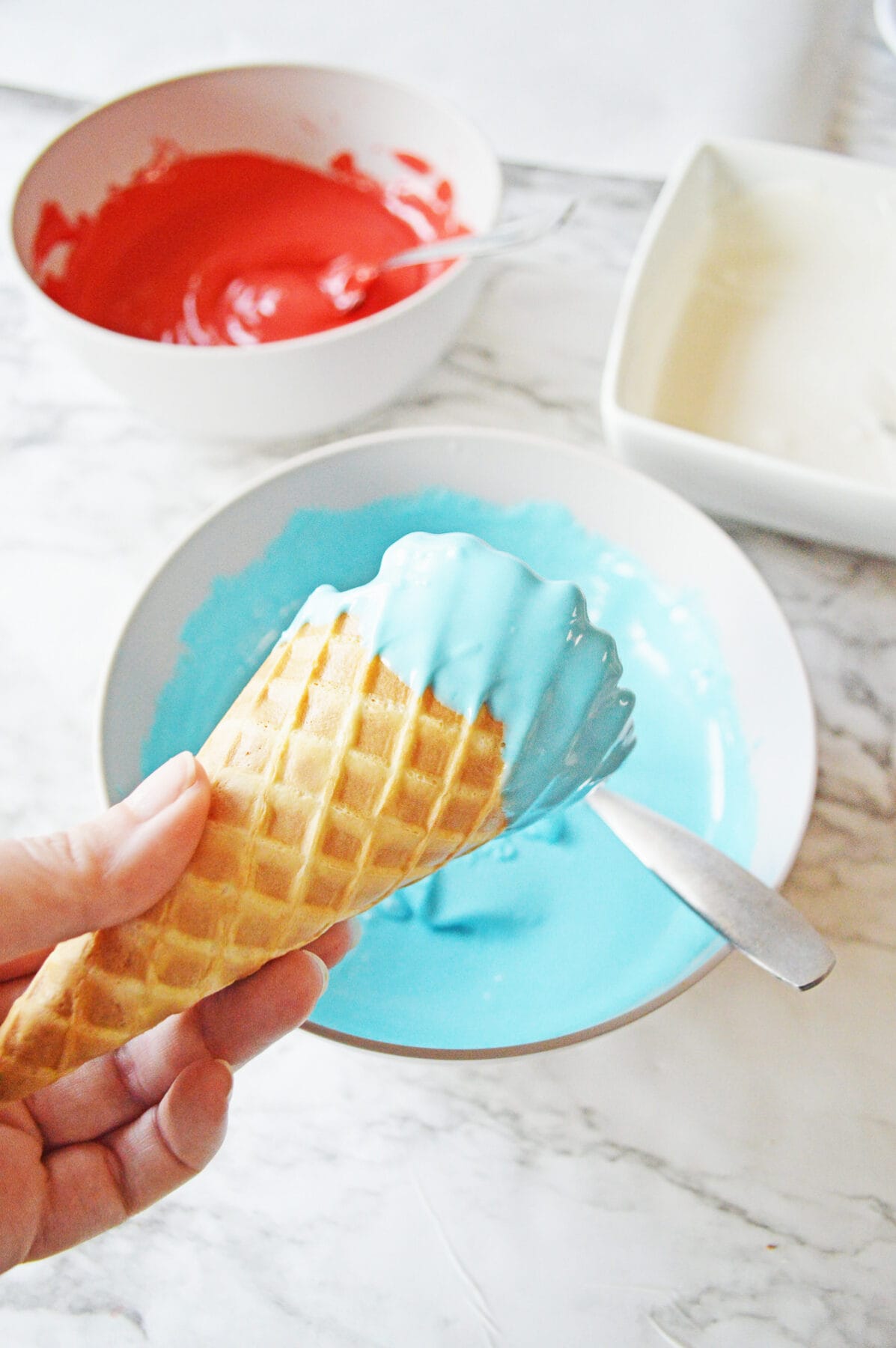 a hand holding a waffle cone covered on the rim with blue chocolate over a bowl with melted blue chocolate.