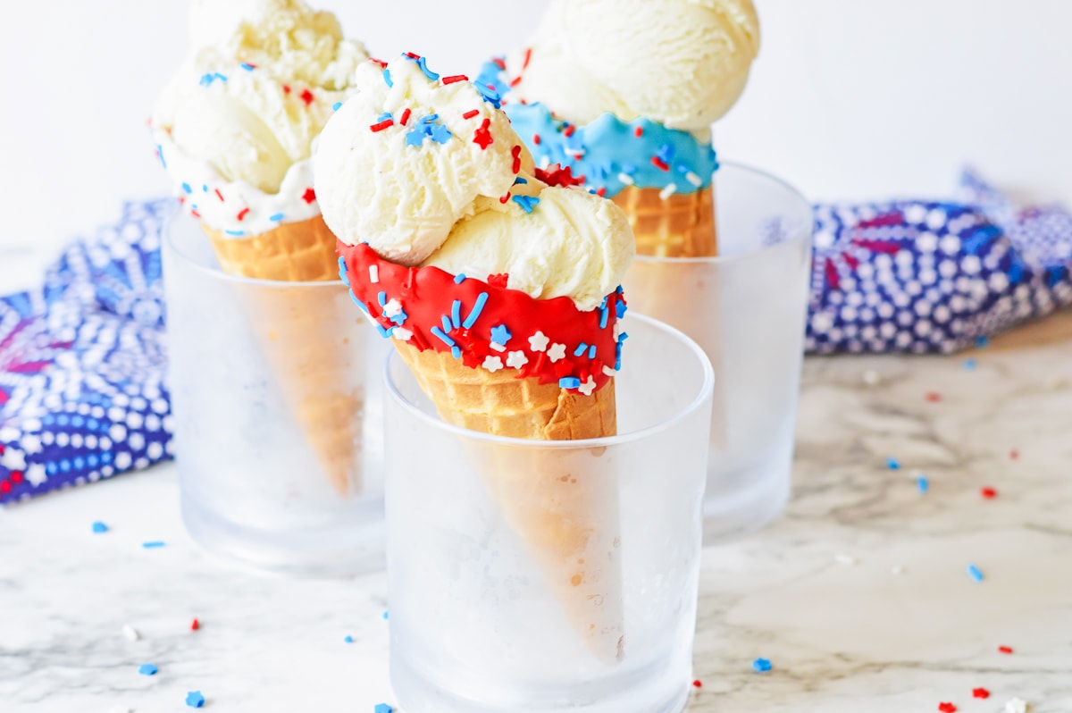 3 fourth of july themed chocolate dipped cones in cups with sprinkles and ice cream.