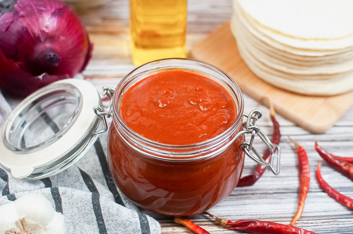 red enchilada sauce in a glass jar with lid on a striped napkin with red chilis to the right.