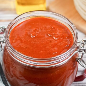 close up of red enchilada sauce in a mason jar with chilis to the side and stack of tortillas in the background.