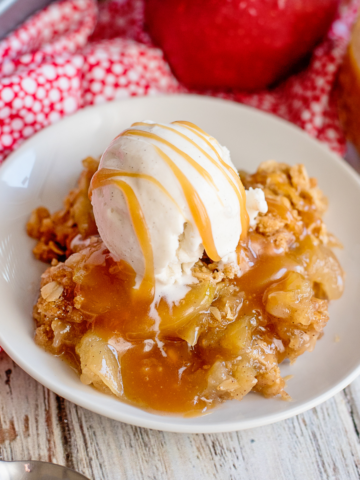 apple crisp with a scoop of vanilla ice cream and a drizzle of caramel sauce on a white plate with apple in the background.