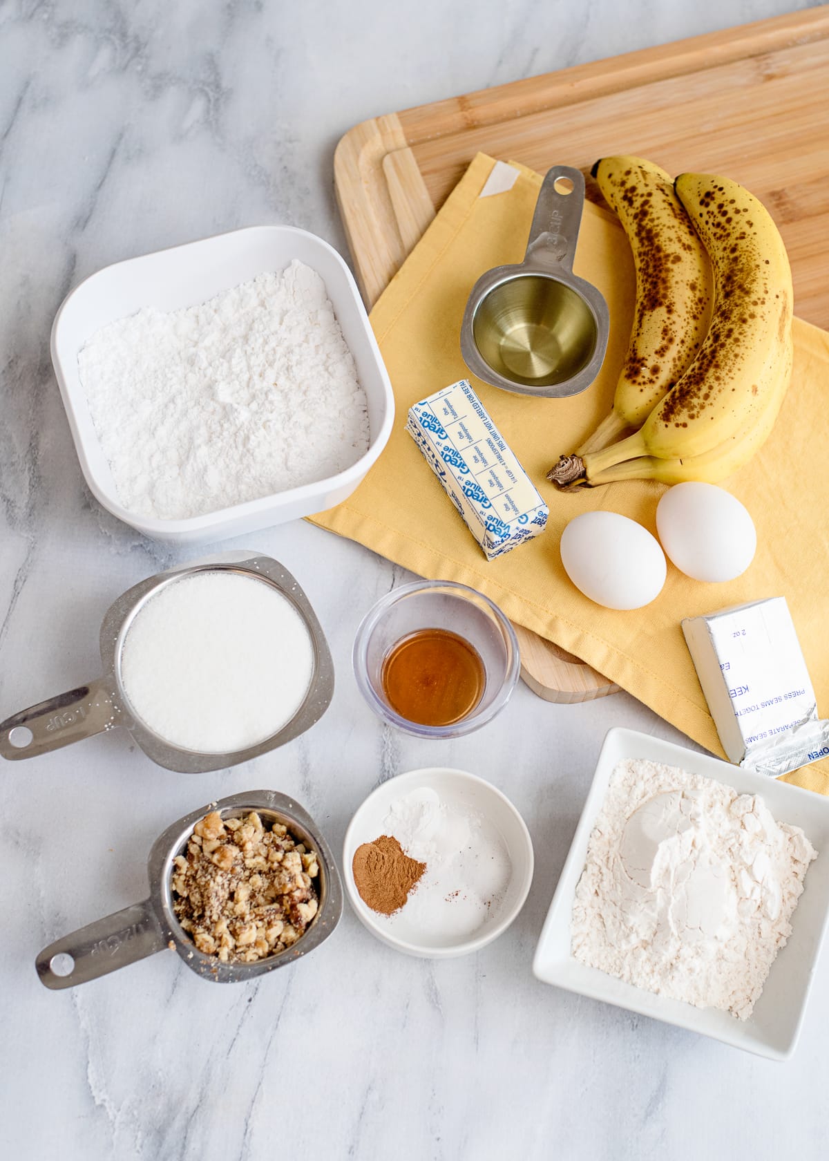 cream cheese, powdered sugar, bananas, eggs, butter, flour, sugar, walnuts, and other ingredients needed for baking a banana nut cake on a marble backdrop with yellow napkin.