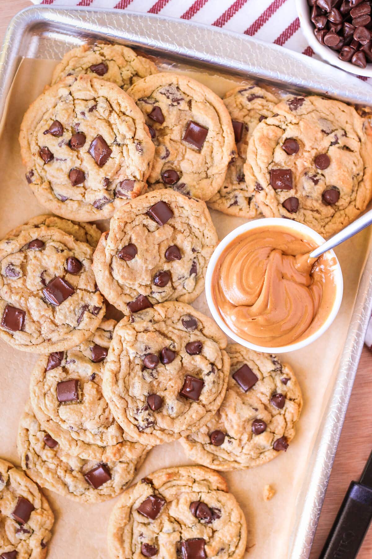 freshly baked peanut butter chocolate chip cookies on a baking sheet with a bowl full of peanut butter next to it with a spoon inside.