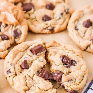peanut butter chocolate chip cookies on a piece of parchment paper and a spoonful of peanut butter in the background, focus is on cookie torn in half slightly.