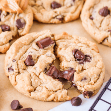 cropped photo of peanut butter chocolate chip cookies on a piece of parchment paper and a spoonful of peanut butter in the background, focus is on cookie torn in half slightly.