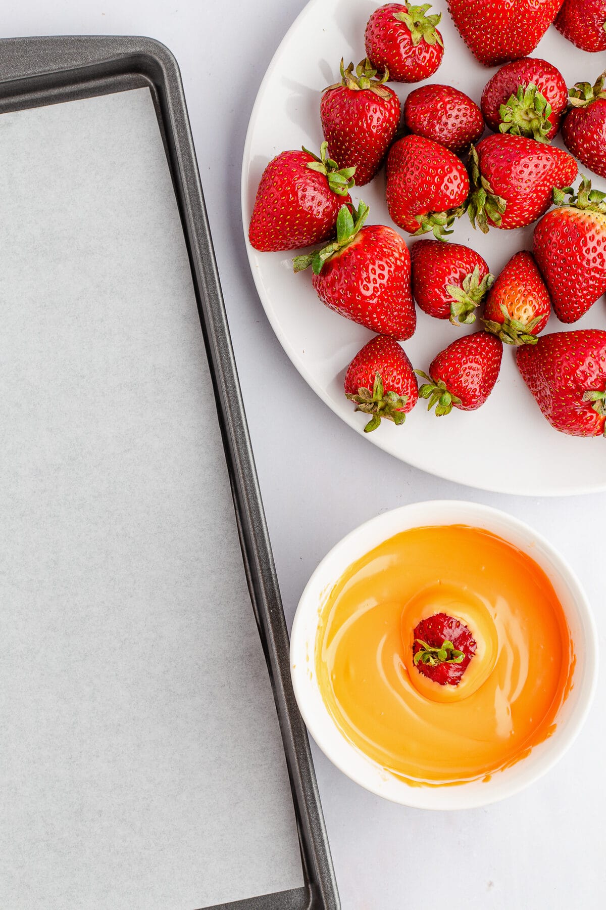 strawberry dipped into a bowl full of orange chocolate with a lined baking sheet to the left and plate of strawberries to the upper right.