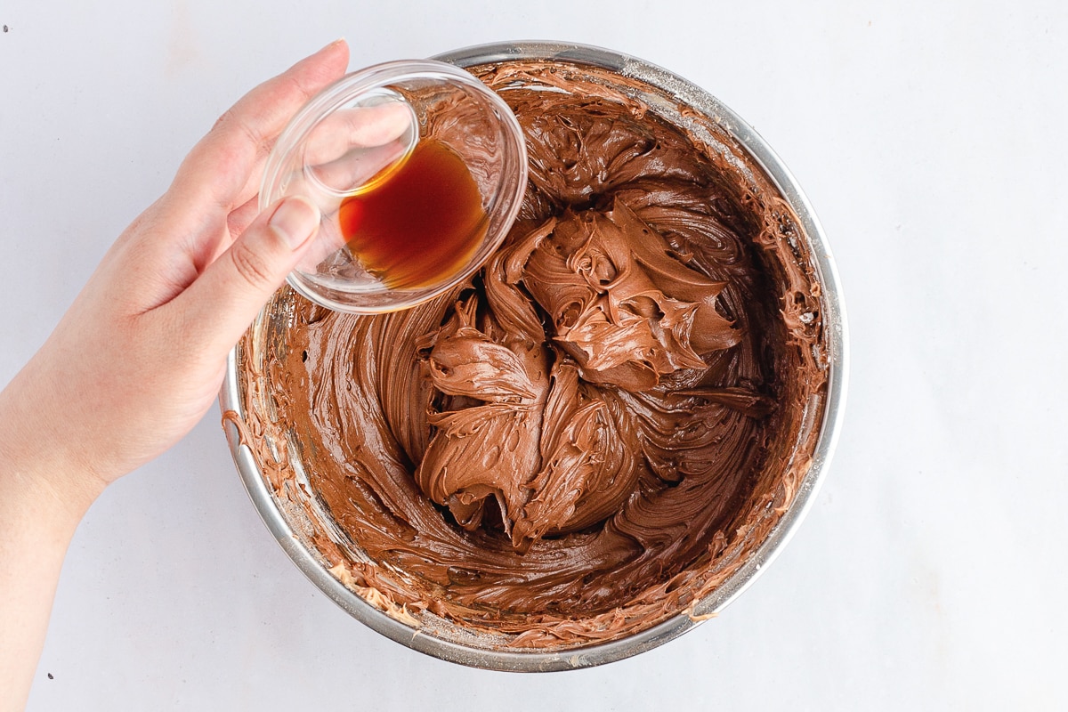 hand holding vanilla extract being added to chocolate frosting in a bowl.