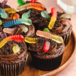 dirt cupcakes with gummy worms on top of them on a wooden plate.