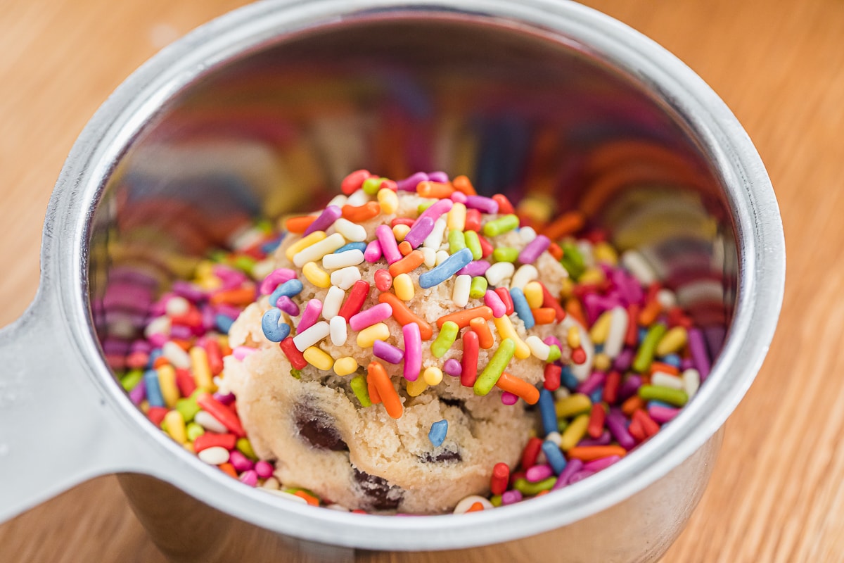 a chocolate chip cookie dough ball covered in sprinkles in a stainless steel bowl with sprinkles.