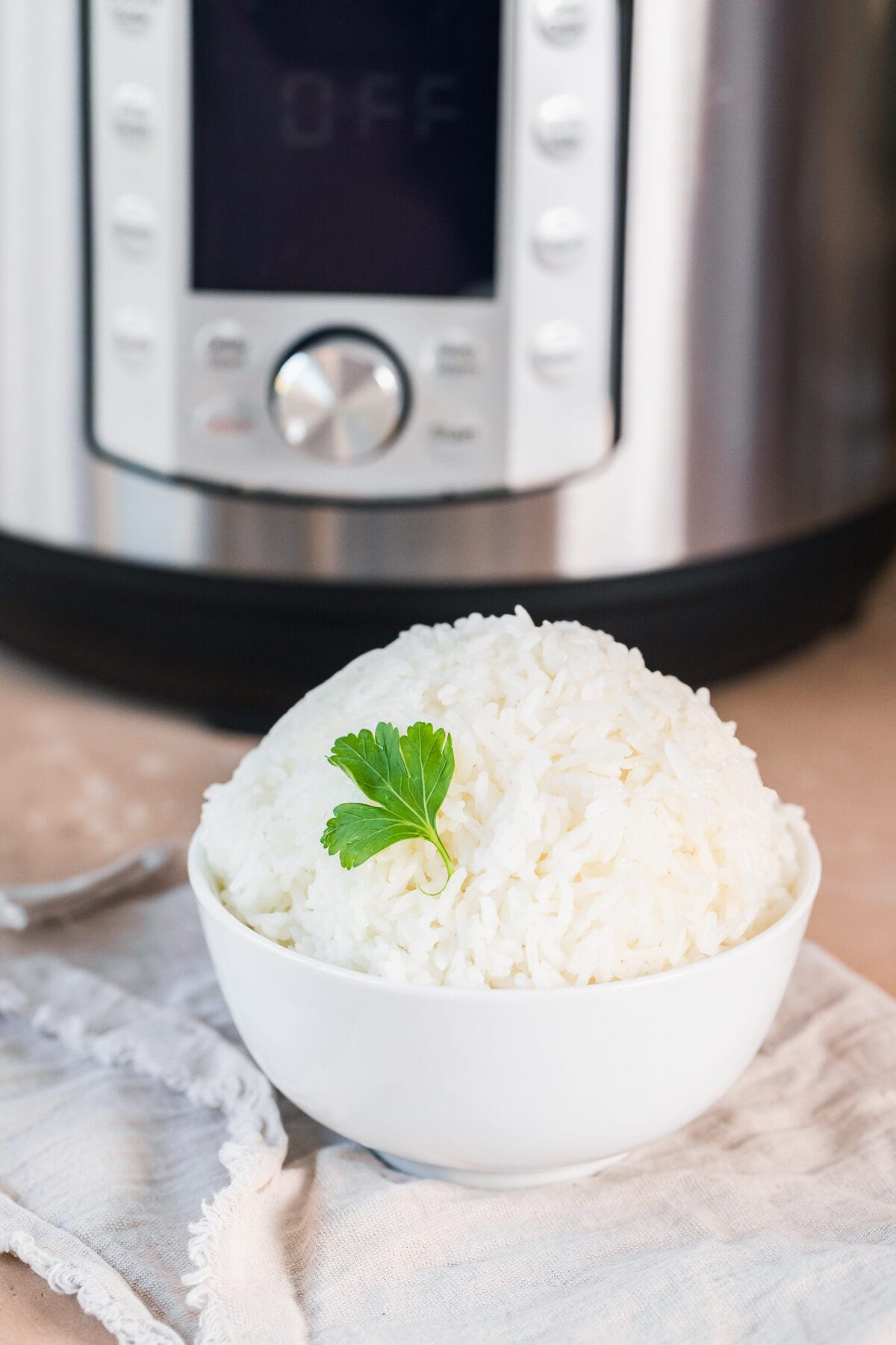 fresh cooked instant pot jasmine rice in a white bowl with a piece of parsley as garnish on a beige napkin in front of an instant pot.