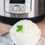 fresh cooked instant pot jasmine rice in a white bowl with a piece of parsley as garnish on a beige napkin in front of an instant pot.