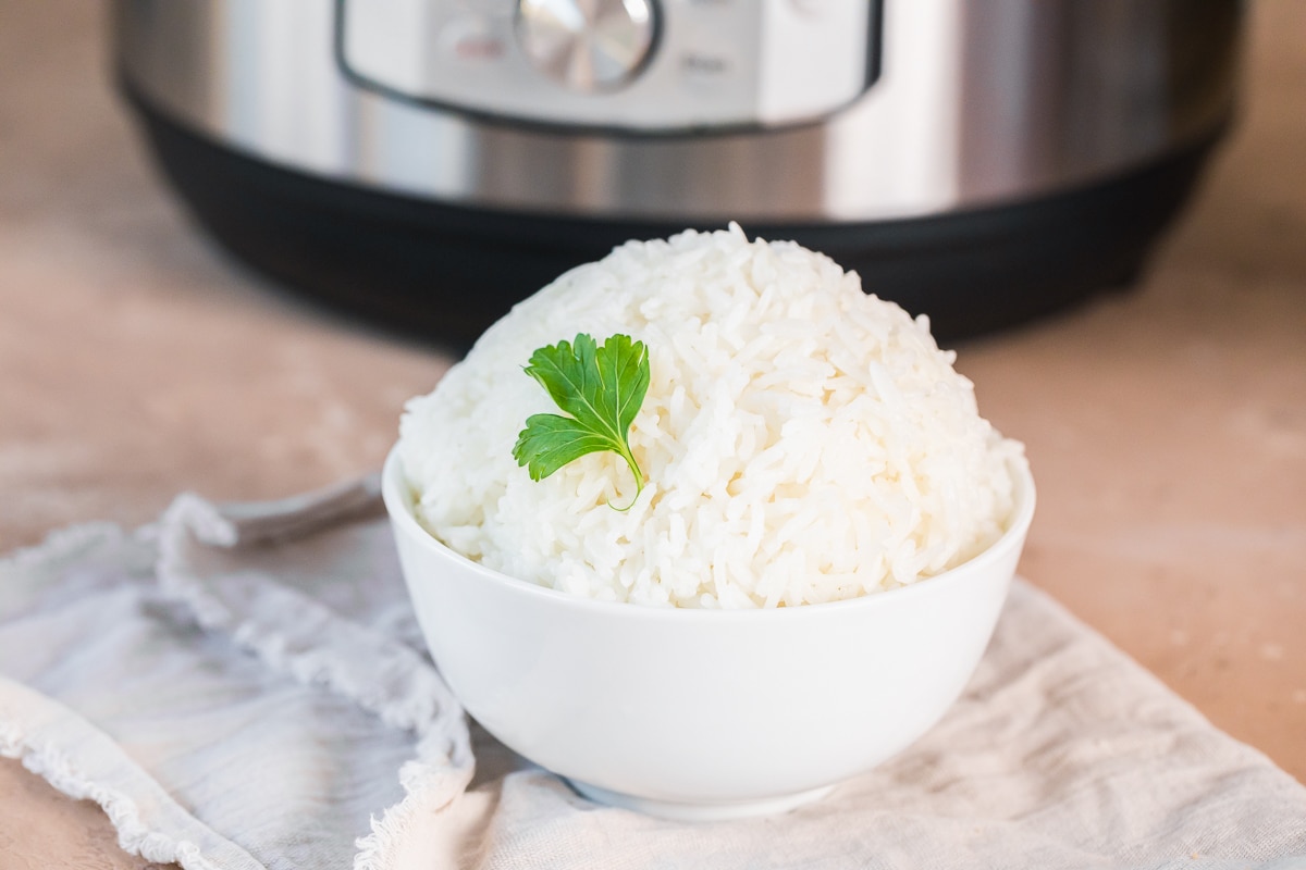 close up of cooked instant pot jasmine rice in a white bowl with a piece of parsley as garnish on a beige napkin in front of an instant pot.