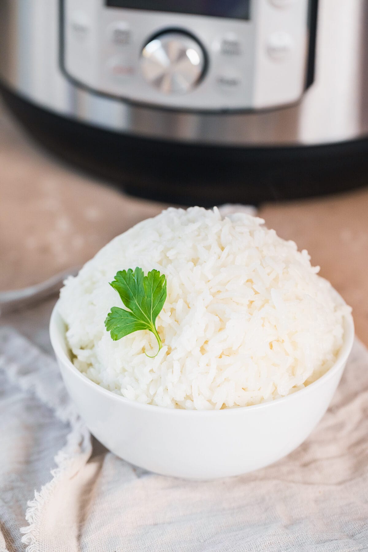 cooked instant pot jasmine rice in a white bowl with a piece of parsley as garnish on a beige napkin in front of an instant pot.