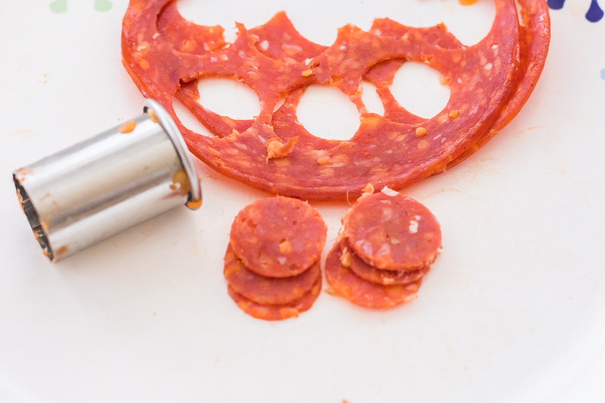 small round cutter with pieces of mini pepperoni next to it and slices of pepperoni with holes in it from cutting mini pepperoni slices.
