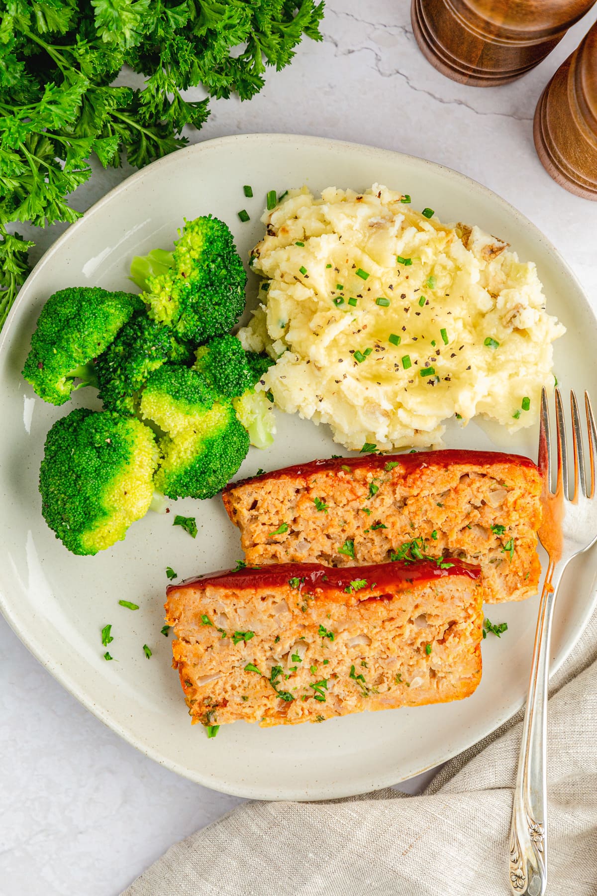 2 slices of chicken meat loaf on a plate with broccoli, garlic mashed potatoes and a spoon and fork.